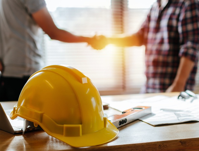 yellow safety helmet on workplace desk with construction worker team hands shaking greeting start up plan new project contract in office center at construction site, partnership and contractor concept
